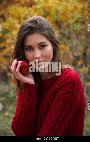 Picknick im Herbstwald. Eine schöne asiatische Frau in einem roten Pullover sitzt auf einem Karos neben einem Korb mit Thermoskannen, Äpfeln und Birnen Stockfoto