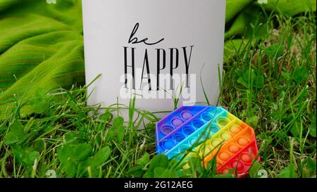 Neue beliebte Silikon bunte Regenbogen Antistress Pop es Spielzeug für Kind auf dem Gras in der Nähe der Geschenkbox mit der Aufschrift Be happy. Speicherplatz kopieren. Einfache Grübchen Stockfoto