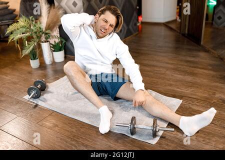 Erschöpfter, aktiver Millennials-Mann, der zu Hause auf einer Fitnessmatte sitzt, sich nach dem Training ausruhen, müde getönter junger Mann in Sportkleidung entspannen sich beim Trainieren Stockfoto