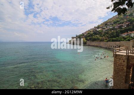 Der historische Kızıl Kule (Roter Turm), Burgmauer und Ruinen des historischen Hafens von Alanya, Menschen, die im Meer baden und am Strand spazieren gehen. Stockfoto