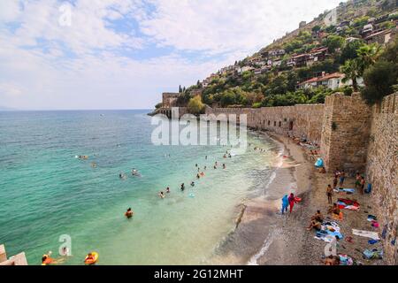 Der historische Kızıl Kule (Roter Turm), Burgmauer und Ruinen des historischen Hafens von Alanya, Menschen, die im Meer baden und am Strand spazieren gehen. Stockfoto
