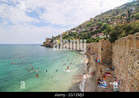 Der historische Kızıl Kule (Roter Turm), Burgmauer und Ruinen des historischen Hafens von Alanya, Menschen, die im Meer baden und am Strand spazieren gehen. Stockfoto