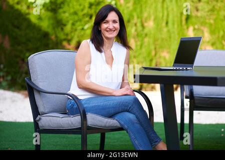Porträt einer Frau, die auf die Kamera schaut, die im Garten ihres Hauses neben einem Laptop sitzt Stockfoto