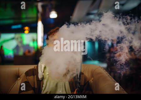 Mann im Restaurant raucht Shisha. Starker Rauch von Shisha. Junger Mann auf dem Sofa im Café. Großer Fernsehbildschirm im Hintergrund. Entspannen Sie sich in der Shisha Bar. Stockfoto