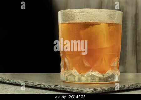 Ein Glas mit einem alkoholischen Getränk und Eis. Scotch Klebeband auf einem Steinständer mit Copyspace. Whiskey mit Eis. Hintergrundbeleuchteter Hintergrund und ein Glas starken, alco Stockfoto