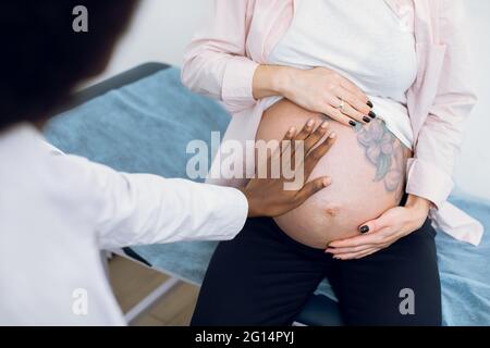 Beschnittenes Nahaufnahme-Bild einer nicht erkennbaren schwarzen Ärztin, die im Krankenhaus eine junge Schwangere untersucht, die auf der Couch sitzt. Cute schwanger Bauch mit Blumen Tattoo Stockfoto