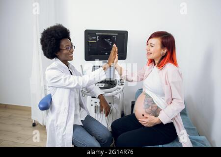 Glückliche, attraktive, schwanger-kaukasische Patientin, die ihrer lächelnden afrikanisch-weiblichen Geburtshelferin eine hohe fünf gab und über eine Schwangerschaft nach Ultraschalluntersuchungen sprach Stockfoto