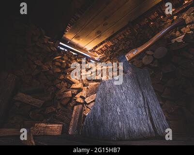 Holzschuppen mit einer bedrohlichen Axt und einem großen Holzstamm mit viel gestapeltem Brennholz im Hintergrund. Lichteinfall im Holzschuppen-Nahaufnahme. Stockfoto