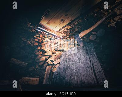 Holzschuppen mit einer bedrohlichen Axt und einem großen Holzstamm mit viel gestapeltem Brennholz im Hintergrund. Lichteinfall im Holzschuppen-Nahaufnahme. Stockfoto