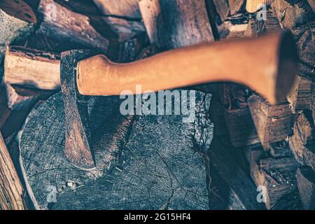 Holzschuppen mit einer bedrohlichen Axt und einem großen Holzstamm mit viel gestapeltem Brennholz im Hintergrund. Lichteinfall im Holzschuppen-Nahaufnahme. Stockfoto