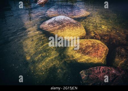 Große Felssteine liegen unter Wasser in einem klaren, transparenten See. Grüner Stein im See unter Wasser, der mit Moos überwuchert ist. Stockfoto