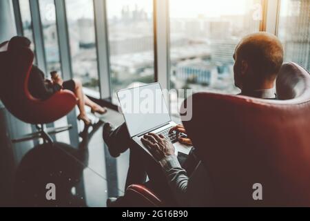 Blick von hinten auf einen Unternehmer, der an seinem Laptop arbeitet, sitzt seine Geschäftspartnerin distanziert in einem roten Sessel, mit einem Papierbecher in ihr Stockfoto