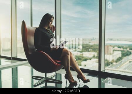 Ein Porträt einer stattlichen und großen Unternehmerin, die auf einem eleganten roten Sessel neben dem Fenster eines modernen Bürohochhauses sitzt und pensiver r Stockfoto