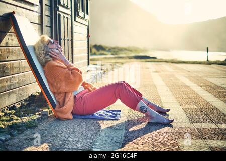 Porträt einer jungen, reifen, blonden kaukasischen Frau im Freien mit einem Bodyboard und einer Sonnenbrille, die mit einem Mobiltelefon spricht. Berria Beach, Kantabrien, Spanien, Stockfoto