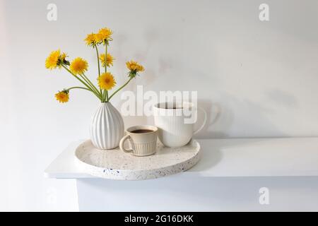 Ein Bouquet von Delelionen in einer weißen, geriffelten Vase und zwei Tassen in verschiedenen Größen mit Kaffee auf einem weißen Tisch. Frühstück. Speicherplatz kopieren Stockfoto