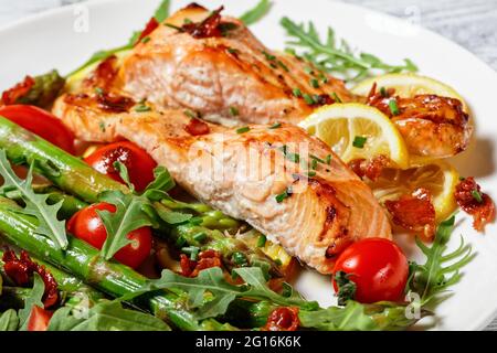 Gegrillte Lachsfilets mit Zitrone und karamellisiertem Speck serviert mit Spargel, Kirschtomaten, wildem Rucola-Salat auf einem weißen Teller Stockfoto