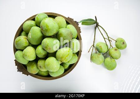 Roher Mango, der traditionell in einem Messinggefäß angeordnet ist, und ein paar grüne Mango, die in der Nähe auf einem weißen strukturierten Hintergrund platziert sind. Stockfoto