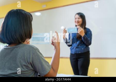 Der asiatische Lehrer schlug vor, den Schülern der Grundschule im Klassenzimmer nach der Quarantäne und Sperrung von Covid-19 die richtige Art und Weise zu empfehlen, eine Maske zu tragen. Stockfoto