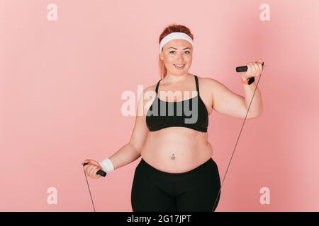 Fröhliche Plus Größe Frau trägt Fitness-Outfit mit Springseil auf rosa Hintergrund stehen. Stockfoto