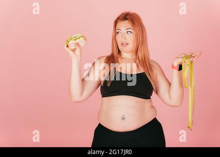 Nachdenkliche Frau mit plus size Körper trägt sportliche Outfit Blick auf Fast Food Burger halten Maßband, während sie auf rosa Studio Hintergrund stehen. Stockfoto