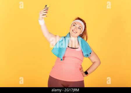 Fröhliche junge Plus-Größe Frau trägt sportliche Outfit und Handtuch nach dem Training hält Smartphone nimmt Selfie, während auf gelbem Hintergrund stehen. Stockfoto