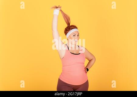 Fröhliche lustige junge Plus-Größe Frau trägt Sport-Outfit hält ihre Pferdeschwanz Frisur lächelnd Blick auf die Kamera in guter Stimmung stehen auf gelbem Rücken Stockfoto