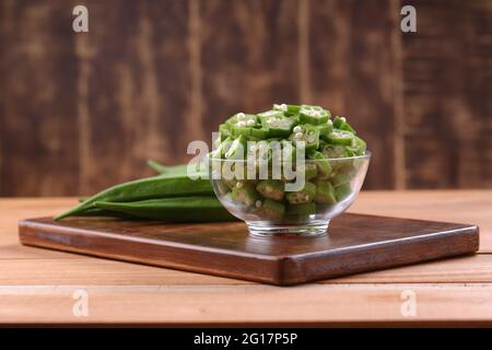 Okra oder Lady's Finger oder Bhindi frisches grünes Gemüse auf einem Holzbrett mit einer Glasschale voller Okra Scheiben Ringe mit Holzhintergrund angeordnet, Stockfoto