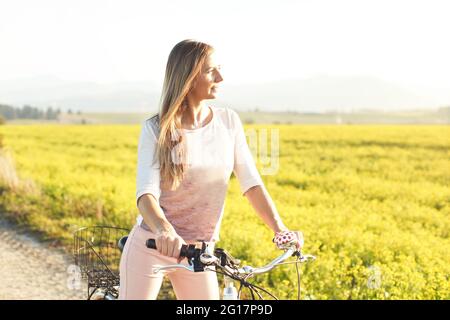 Junge Frau, die neben ihrem Fahrrad über staubige Landstraße steht, starke nachmittägliche Sonnenbeleuchtung im Hintergrund leuchtet auf gelbem Blumenfeld Stockfoto