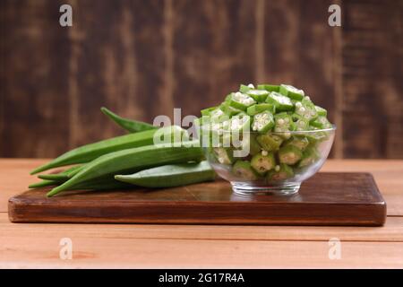 Okra oder Lady's Finger oder Bhindi frisches grünes Gemüse auf einem Holzbrett mit einer Glasschale voller Okra Scheiben Ringe mit Holzhintergrund angeordnet, Stockfoto