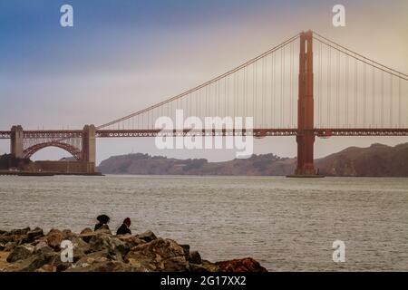 Golden Gate Bridge an einem nebligen Tag und zwei Menschen sitzen an der Bucht von San Francisco Stockfoto