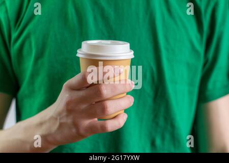 Eine Papiertasse mit Kaffee zum Mitnehmen in der Hand eines jungen Mannes in einem grünen T-Shirt Stockfoto