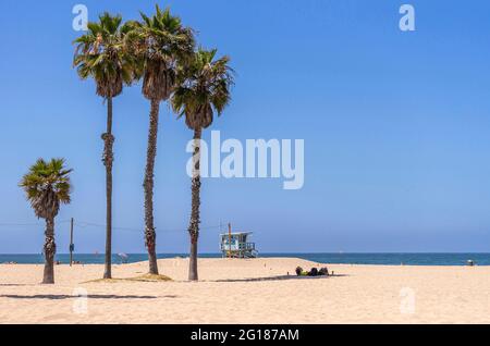 Santa Monica, CA, USA - 20. Juni 2013: Paar ruht am Sandstrand im Palmenschatten unter blauem Himmel. Rettungsschwimmer halten sich hinten in dunkelblauer Weste auf Stockfoto