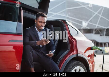 Seriöser afrikanischer Geschäftsmann, der in einem roten Auto mit geöffneter Tür sitzt und sein Smartphone mit Geldgeld in der Hand hält. Luxus Auto wird aufgeladen. Konzept der Einsparungen. Stockfoto