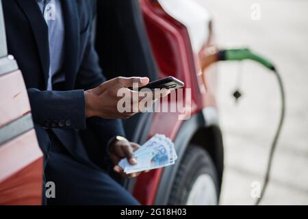 Seriöser afrikanischer Geschäftsmann, der in einem roten Auto mit geöffneter Tür sitzt und sein Smartphone mit Geldgeld in der Hand hält. Luxus Auto wird aufgeladen. Konzept der Einsparungen. Stockfoto