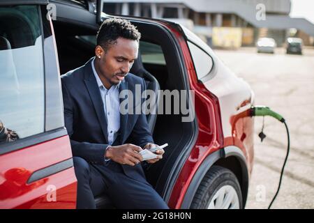 Hübscher afrikanischer Mann im Geschäftsanzug mit Geldkassieren in den Händen, der im Elektroauto sitzt, das lädt. Konzept von Menschen, Transport und Einsparungen. Stockfoto