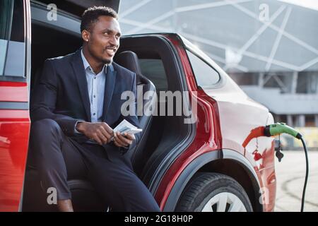 Seitenansicht des glücklichen afrikanisch-amerikanischen Mannes im Geschäftsanzug, der Geld in bar zählt, während er ein Elektroauto lädt. Junger Mann, der im Auto sitzt und sich über das wirtschaftliche Fahrzeug zufrieden fühlt. Stockfoto