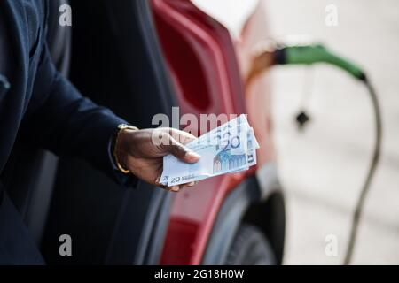 Nahaufnahme der männlichen Hand, die Geld Bargeld über verschwommen Hintergrund des Ladevorgangs Elektroauto hält. Smart african Geschäftsmann im Anzug spart Geld auf ökologische Fahrzeug. Stockfoto