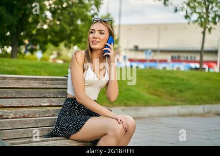 Glückliches Mädchen, das auf dem Handy anruft und auf einer Bank auf der Straße sitzt. Junge blonde Mädchen wartet auf eine Person in einem Park. Hochwertige Fotos Stockfoto