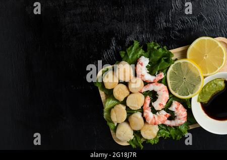 Gebratener Jakobsmuschel mit gekochtem Garnelenschwanz auf einem Salatblatt mit Zitrone, Sojasauce und Wasabi auf dunklem Grund auf einem Holzteller. Selektiver Fokus. Stockfoto