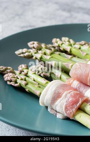 Spargel in Rückenspeck auf einem blaugrünen Teller eingewickelt. Mit einem konkreten Hintergrund Stockfoto