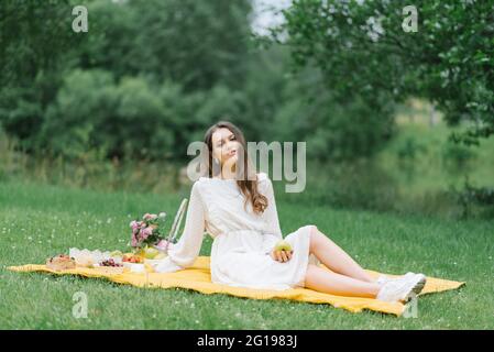 Eine junge schöne Frau in einem weißen Kleid sitzt auf einer Decke auf einem Sommerpicknick auf dem grünen Gras. Schöne Zeit in der Natur im Sommer Stockfoto