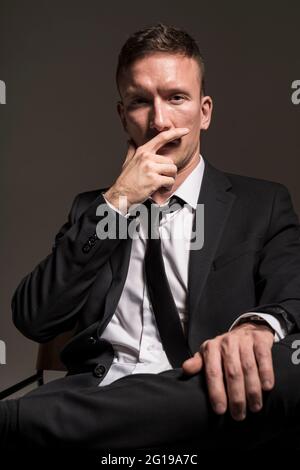 Porträt eines nachdenklichen, selbstbewussten Mannes, in einem Business-Anzug, sitzend mit gekreuzten Beinen vor einem dunklen Hintergrund. Stockfoto