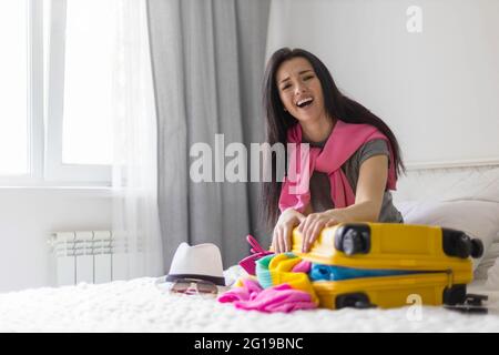 Glückliche asiatische Frau versucht, schließen gelben Koffer voller verschiedener Kleidung Accessoires und Schuhe Stockfoto