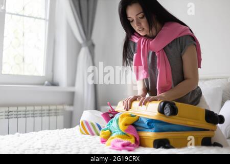 Glückliche asiatische Frau versucht, schließen gelben Koffer voller verschiedener Kleidung Accessoires und Schuhe Stockfoto