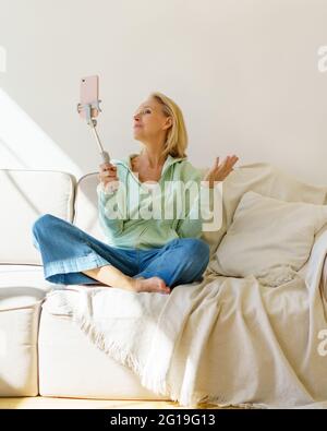Frohes Leben im Ruhestand. Positive ältere Frau, die Selfie auf dem Smartphone macht oder Video-Chat mit der Familie genießt, während sie in Lotuspose auf dem Sofa sitzt und sich zu Hause entspannt. Ältere Frauen mit modernen Technologien Stockfoto