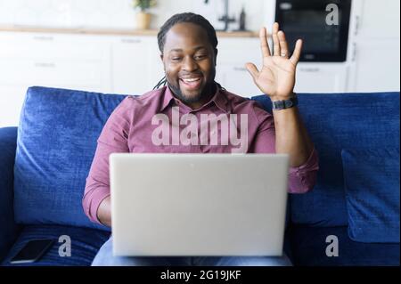 Ein fröhlicher afroamerikanischer Kerl winkt mit der Hand in die Webcam des Laptops, sitzt auf der bequemen Couch und hält virtuelle Meetings ab, macht Videoanruf, bleibt in Kontakt mit Freunden oder der Familie Stockfoto