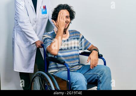 Gutaussehender hispanischer Mann, der auf dem Rollstuhl sitzt und einen Halsband trägt, der ein Auge mit der Hand bedeckt, selbstbewusstes Lächeln im Gesicht und überraschende Emotionen. Stockfoto