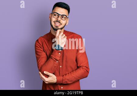 Junger hispanischer Mann mit Bart, Geschäftsshirt und Brille mit Hand am Kinn, der über Fragen nachdenkt, nachdenklichen Ausdruck. Lächelnd mit thoughtfu Stockfoto