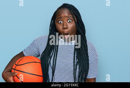 Junge schwarze Frau mit Zöpfen hält Basketball-Ball verängstigt und erstaunt mit offenem Mund für Überraschung, Unglauben Gesicht Stockfoto