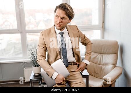 Ein gutaussehender, nachdenklicher kaukasischer Geschäftsmann, der sich mit einem Laptop in den Händen auf den Tisch lehnte und wegschaute. Junge ernsthafte nachdenkliche CEO Unternehmer denken an Stockfoto
